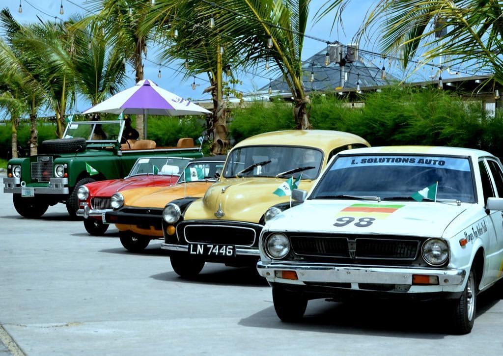 A row of classic cars decorated with Nigerian flags, from a vintage Land Rover to a white rally car numbered 98